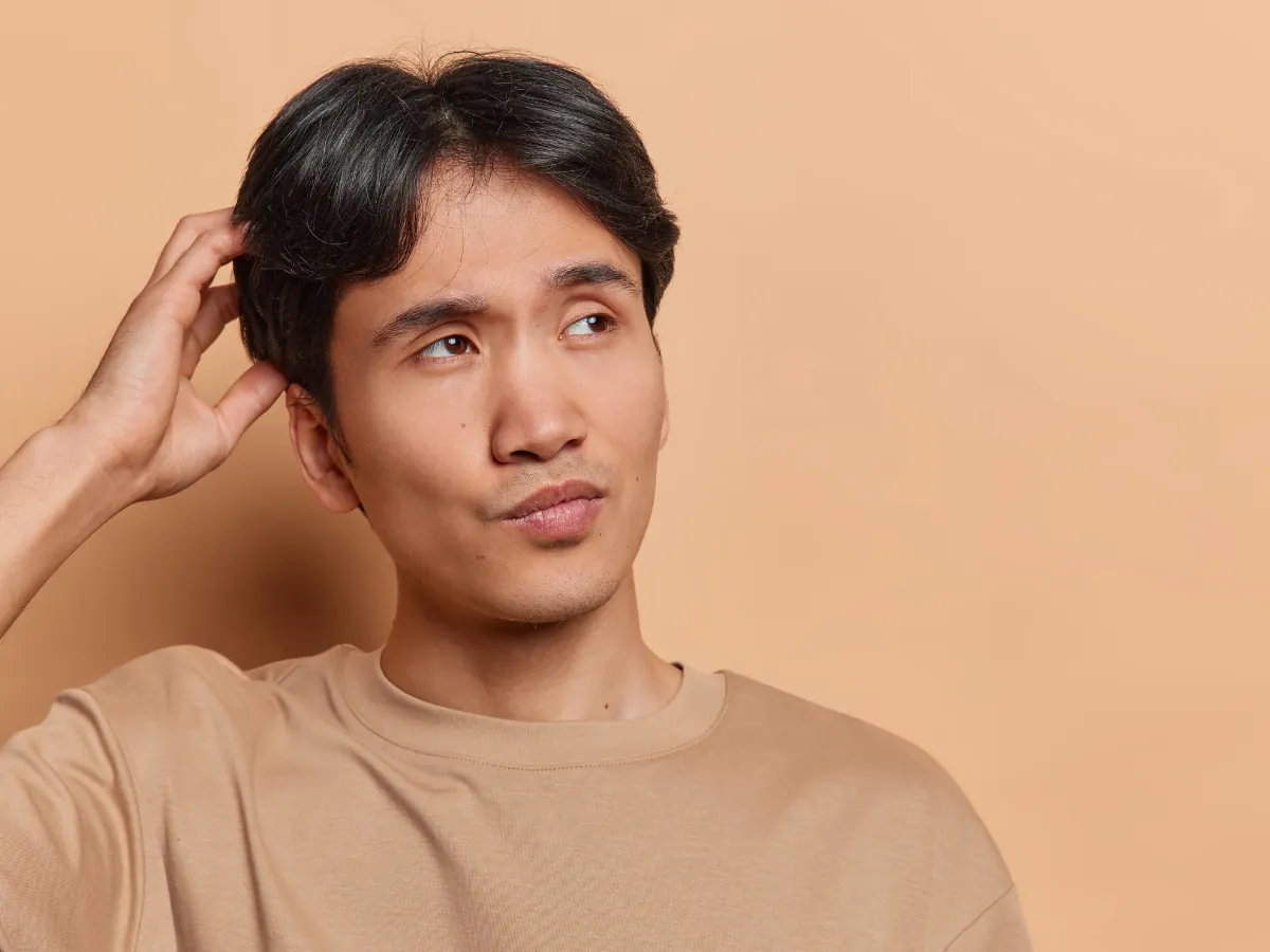 A confused young Filipino man scratching his head, looking uncertain while facing sideways against a beige background.